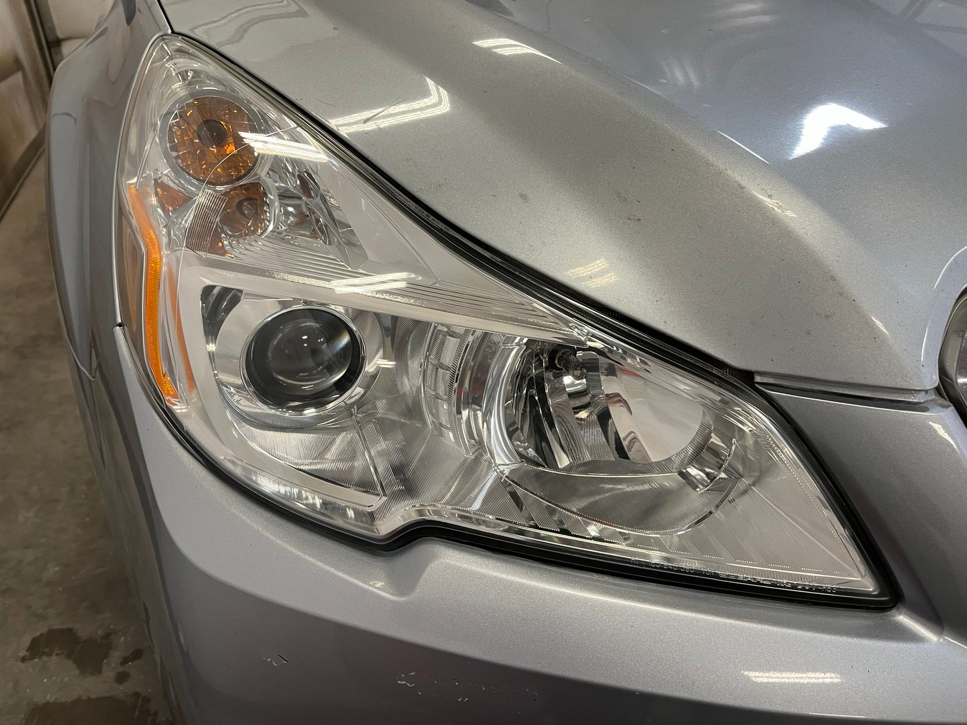 Close-up of a silver car’s front headlight and hood, showing chrome details and an amber turn signal.