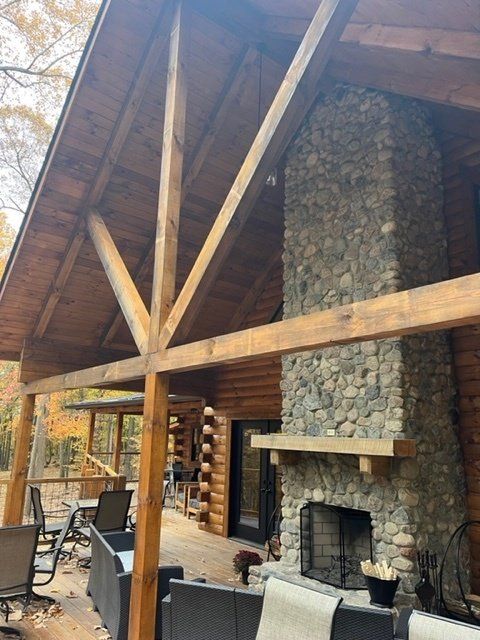 Wooden deck with log cabin and stone fireplace. Exposed wooden beams support the roof. Outdoor seating.