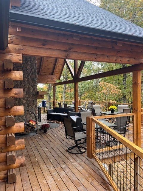 Covered wooden deck with outdoor furniture, overlooking a wooded area. Log cabin visible.