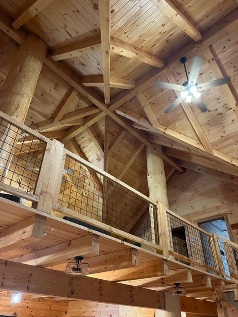 Interior view of a rustic wooden cabin with a loft and high ceilings, showing exposed beams, natural wood, and a ceiling fan.