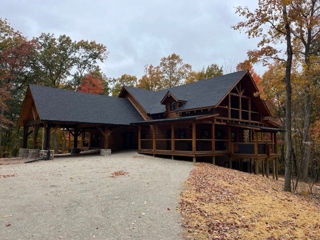 Wooden cabin with porch and carport, gravel driveway, surrounded by fall foliage.