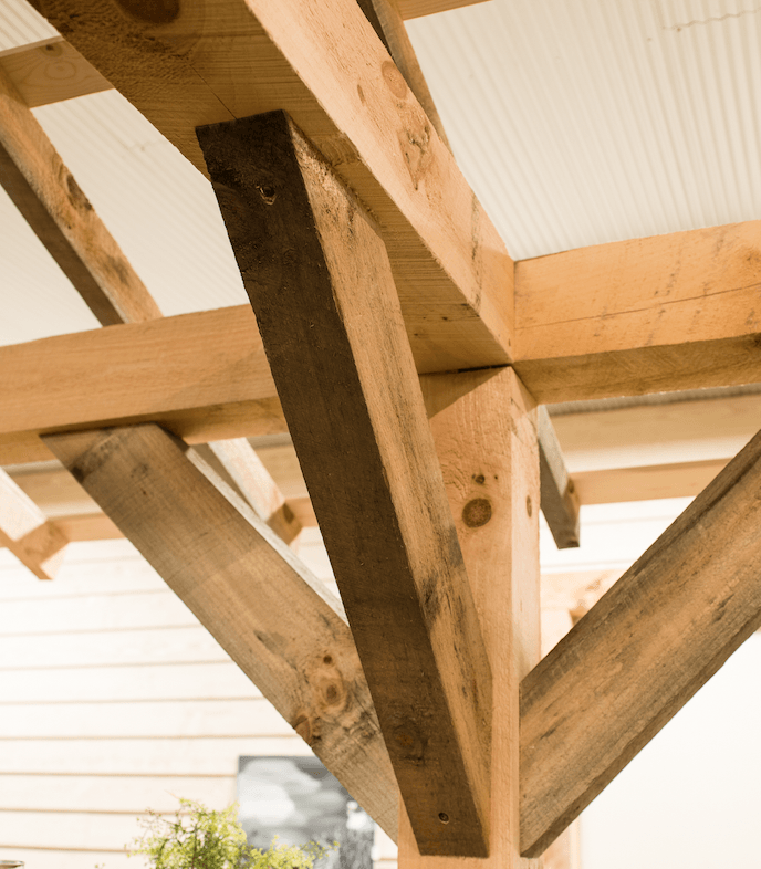 Wooden pergola structure with diagonal bracing, natural wood color, and a white corrugated roof.