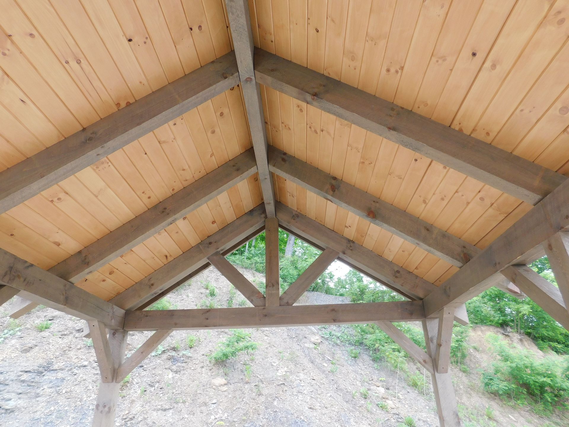 Wooden roof frame with natural wood planks and gray beams, against a green hillside backdrop.