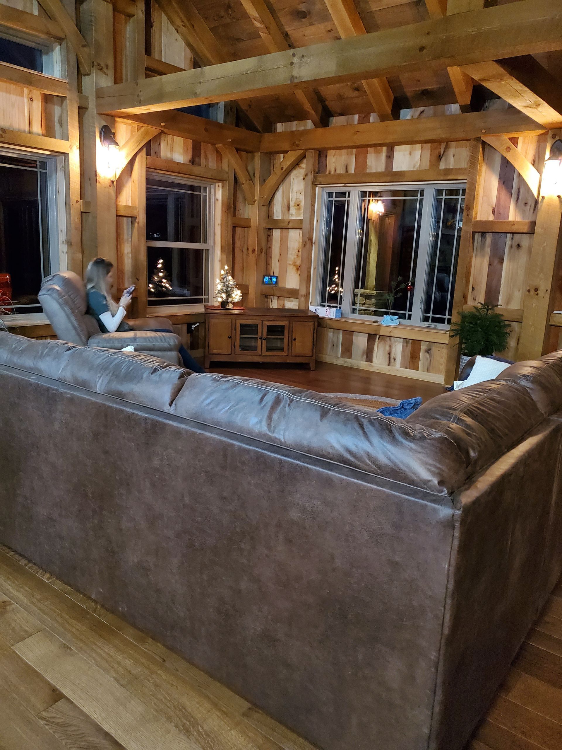 Living room with exposed wood beams, leather sectional, windows, and a person on a couch.