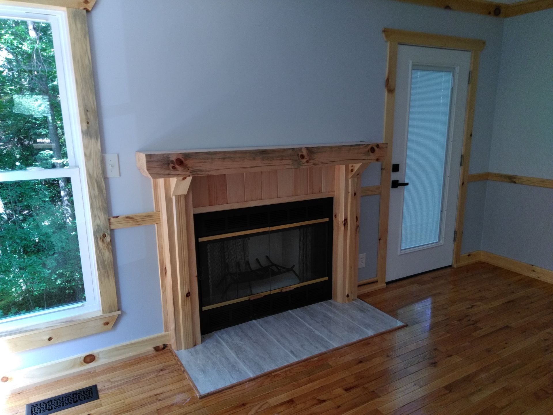 Fireplace with wooden mantel, light gray walls, window and door with wooden trim, hardwood floors.