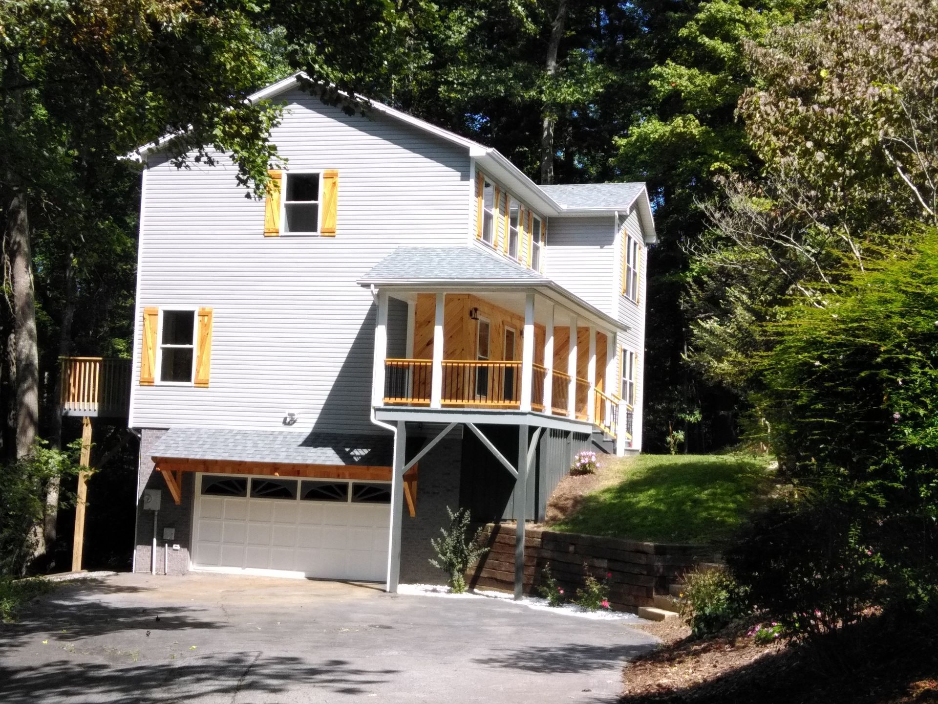 Two-story gray house with garage, porch, and yellow shutters set among trees on a sunny day.