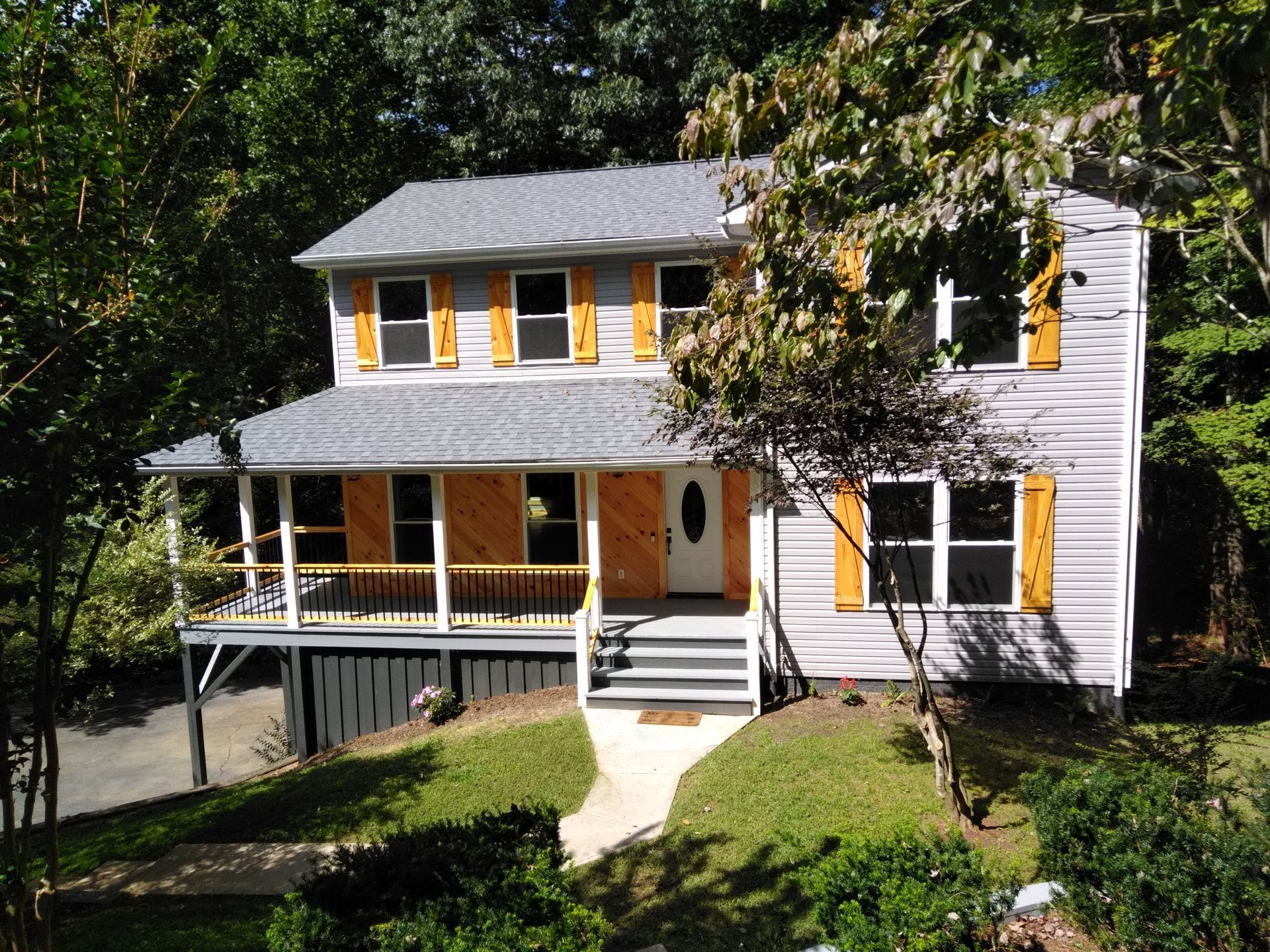 Two-story house with gray siding, gray roof, wooden shutters, and a front porch, surrounded by greenery.