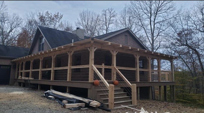 Wooden porch with railings and steps on a house with a dark roof, surrounded by trees.