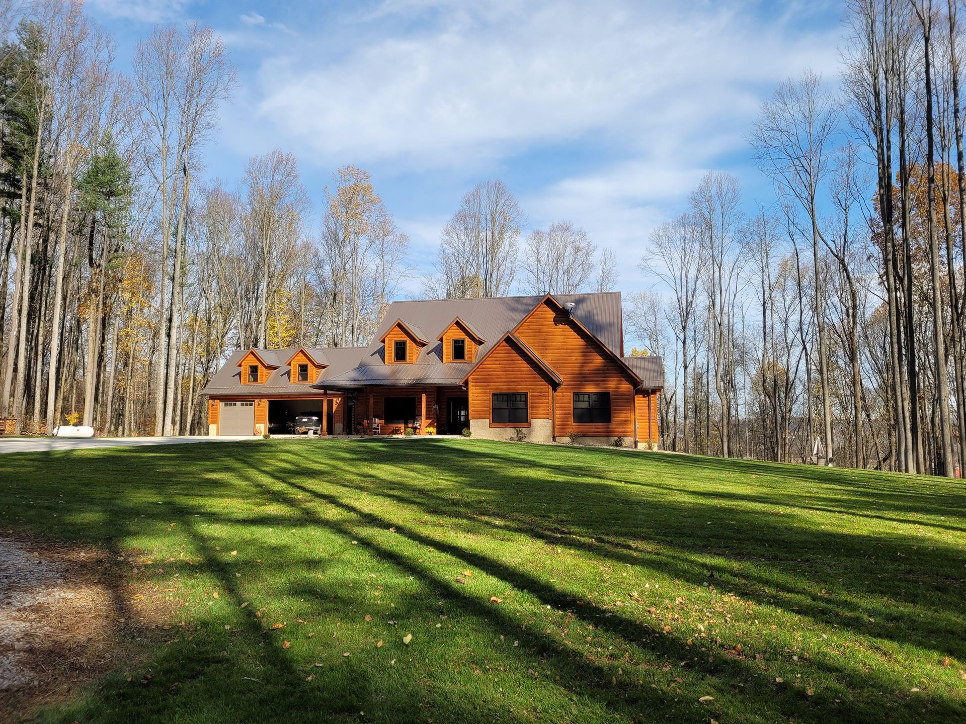 Brown log cabin home on a grassy hill, surrounded by trees under a blue sky.