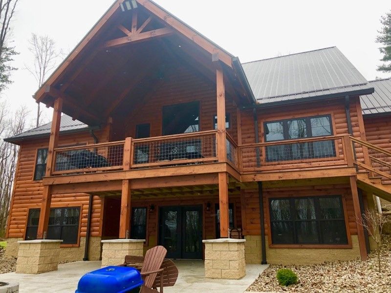 Wooden cabin-style house with a covered porch and deck. Metal roof, stone pillars, and a patio with outdoor furniture.