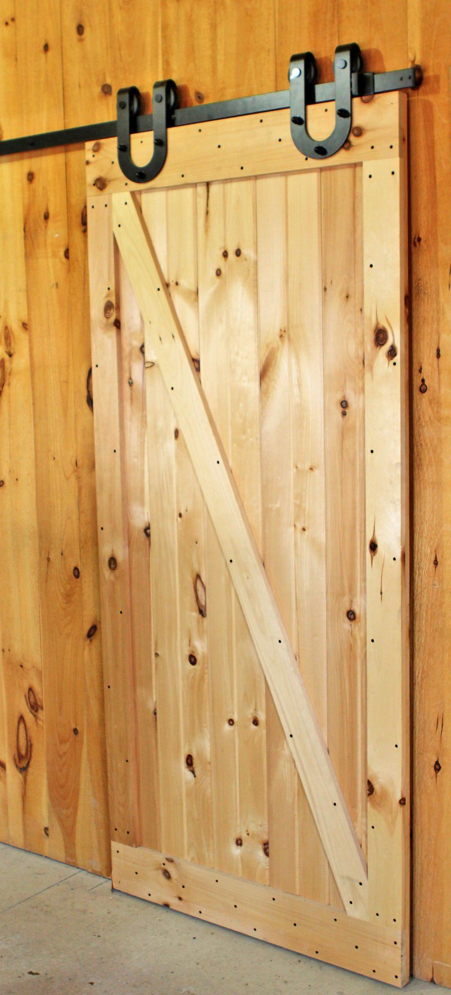 Barn door, light wood, with black hardware. Angled brace across the face. Mounted on wood siding.