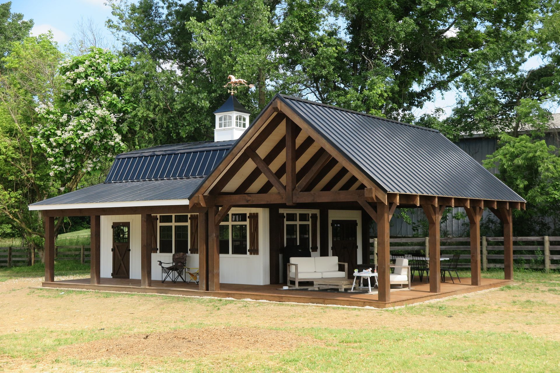 Barn-style building with black roof, brown wooden porch, white walls, and a weathervane. Set in a grassy area with trees.