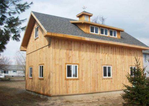 Wooden barn with a gray roof and small windows, built on a concrete foundation in a yard.