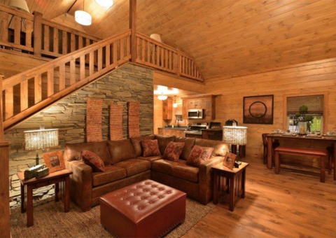 Cozy cabin living room with leather sectional, stone fireplace, and wooden staircase.