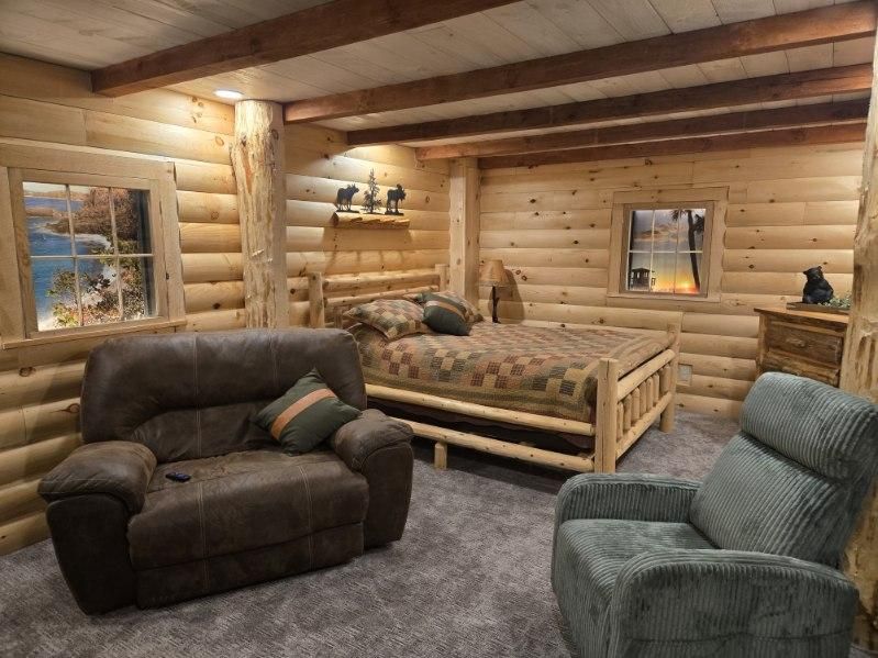 Log cabin bedroom with log walls, wooden bed, two recliners, and landscape art windows.