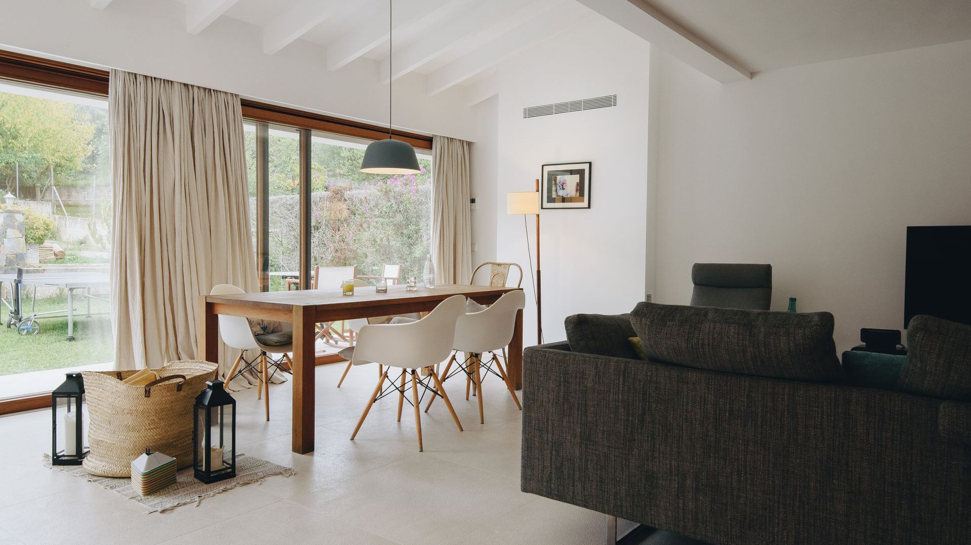 Comedor luminoso con mesa de madera, sillas blancas, lámpara colgante y grandes ventanales con vistas a un jardín verde.