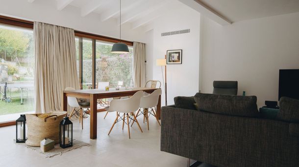 Comedor luminoso con mesa de madera, sillas blancas, lámpara colgante y grandes ventanales con vistas a un jardín verde.