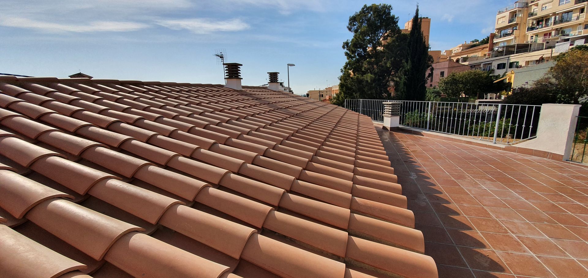 Tejado de tejas de terracota con una terraza soleada y árboles a lo lejos bajo un cielo despejado.