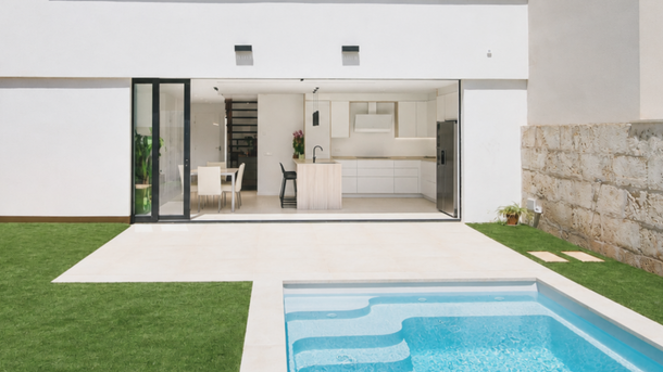 Patio interior de una casa moderna de color blanco con puertas de cristal, césped, muro de piedra y una pequeña piscina azul.