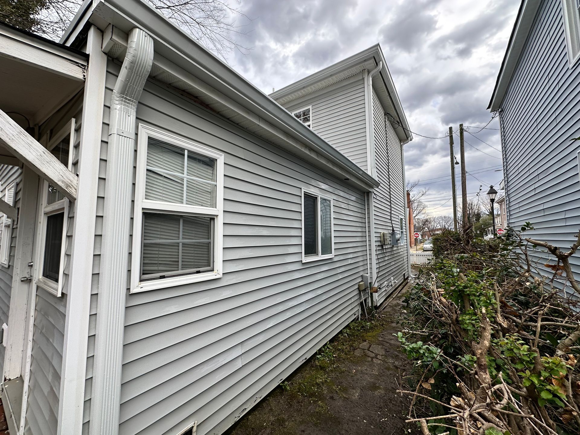 A side view of a light gray, vinyl-sided house with white trim and windows, situated next to a narrow path and bushes.