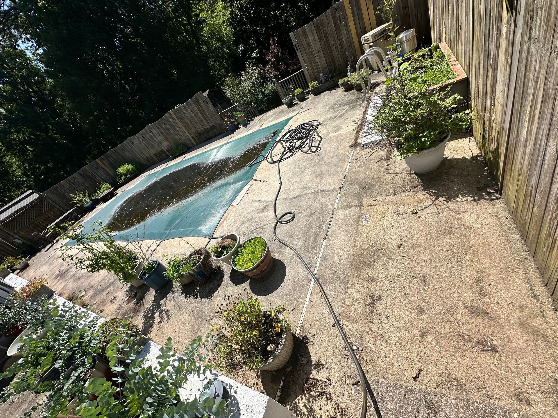 A covered swimming pool with debris on the surface, surrounded by a stone patio, potted plants, and a wooden fence.