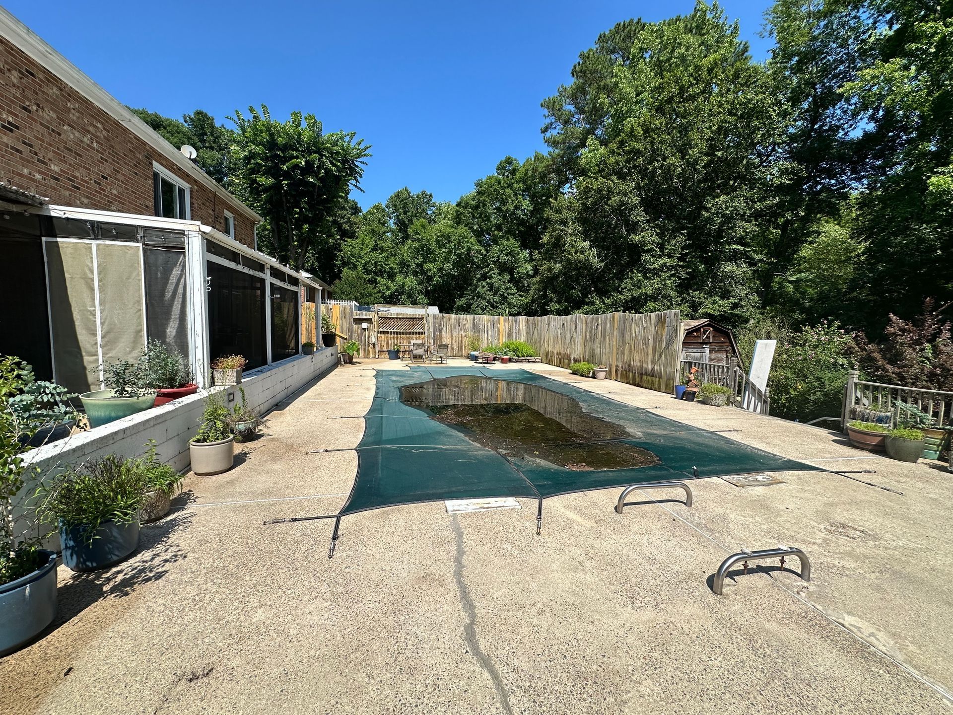 A backyard with a covered swimming pool, patio area, brick house exterior, and wooden fence surrounded by green trees.