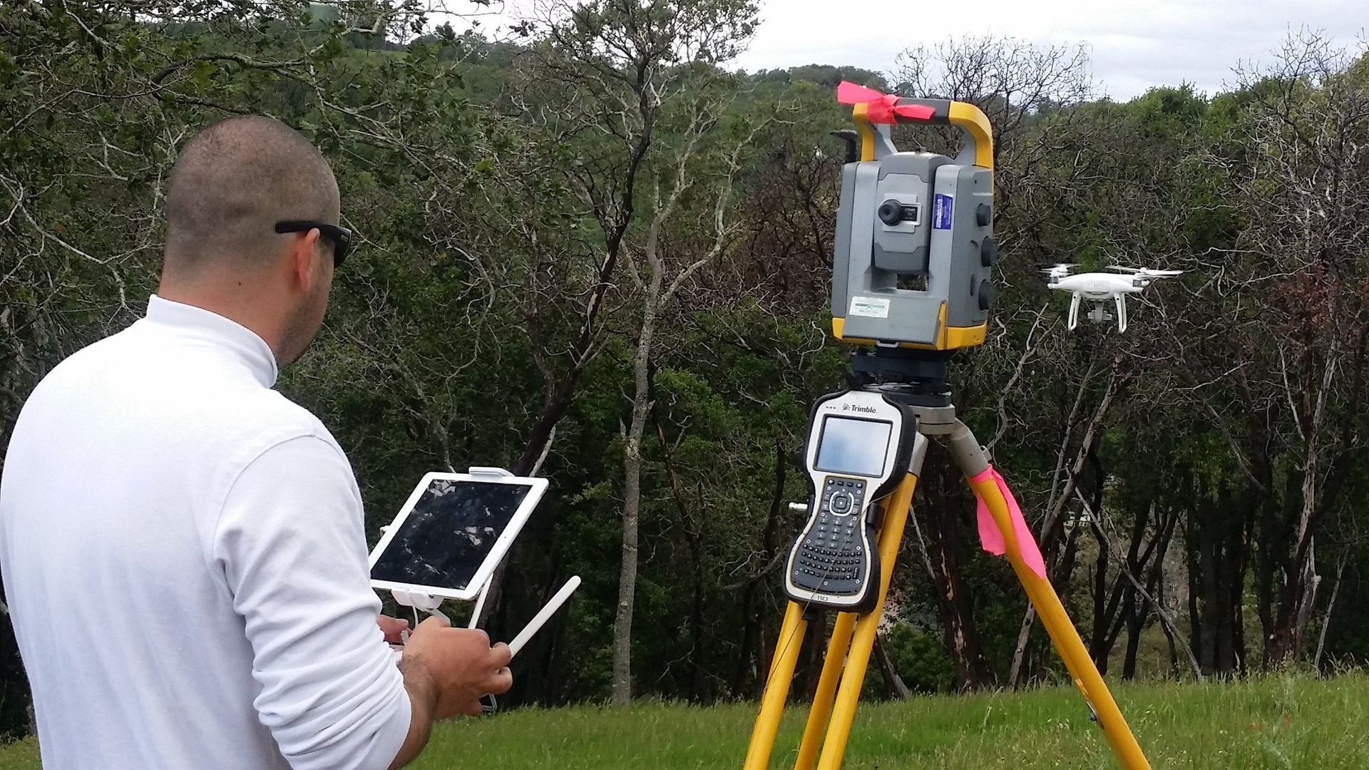Man operating surveying equipment in a wooded area. He holds a tablet, controlling a device on a tripod.