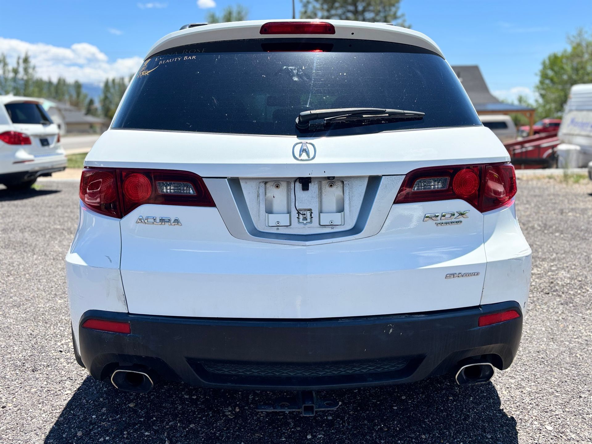 Rear view of a white Acura SUV parked on a gravel lot under a blue sky.