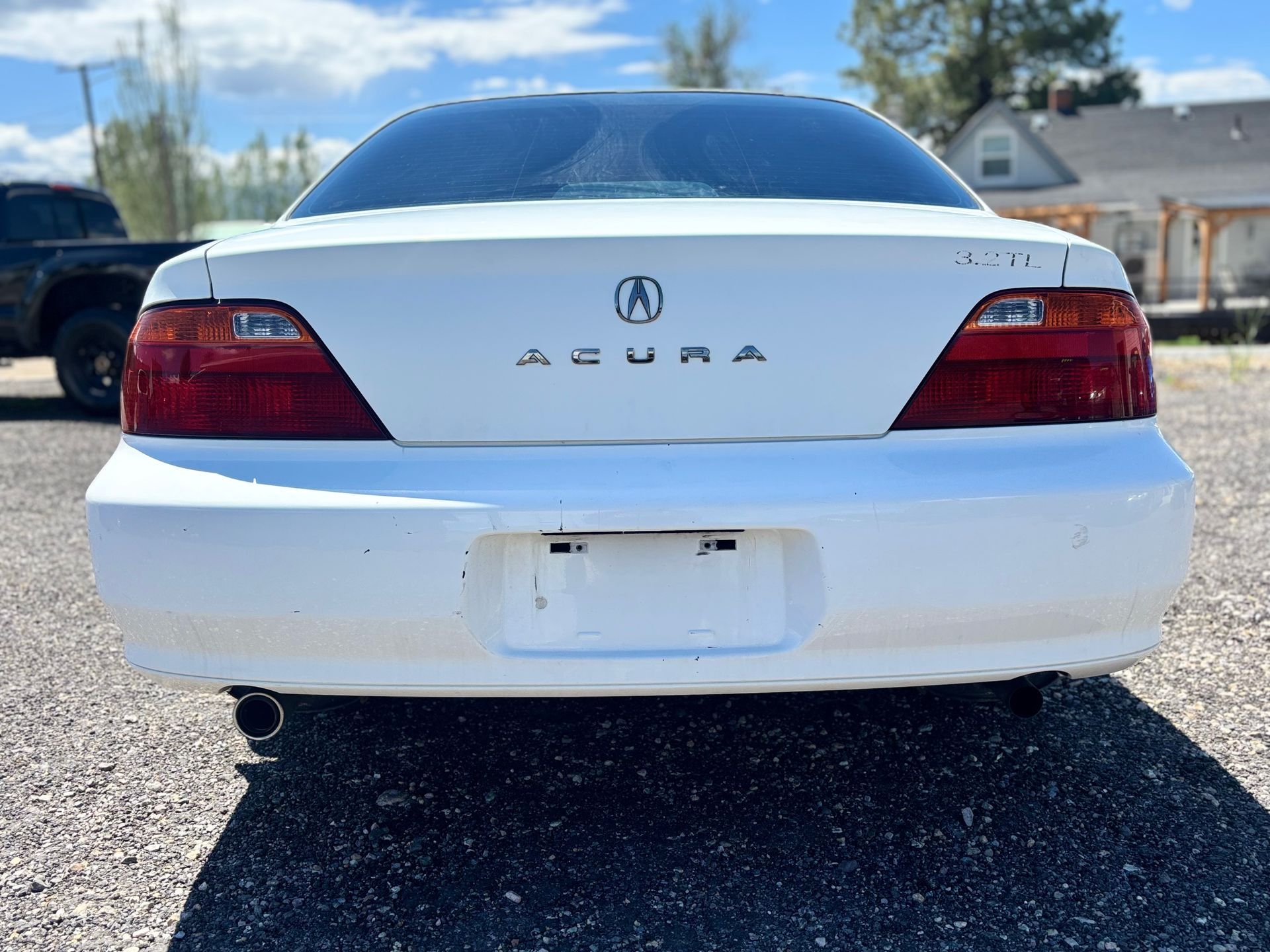 Rear view of a white Acura sedan parked on a gravel lot under a clear sky.