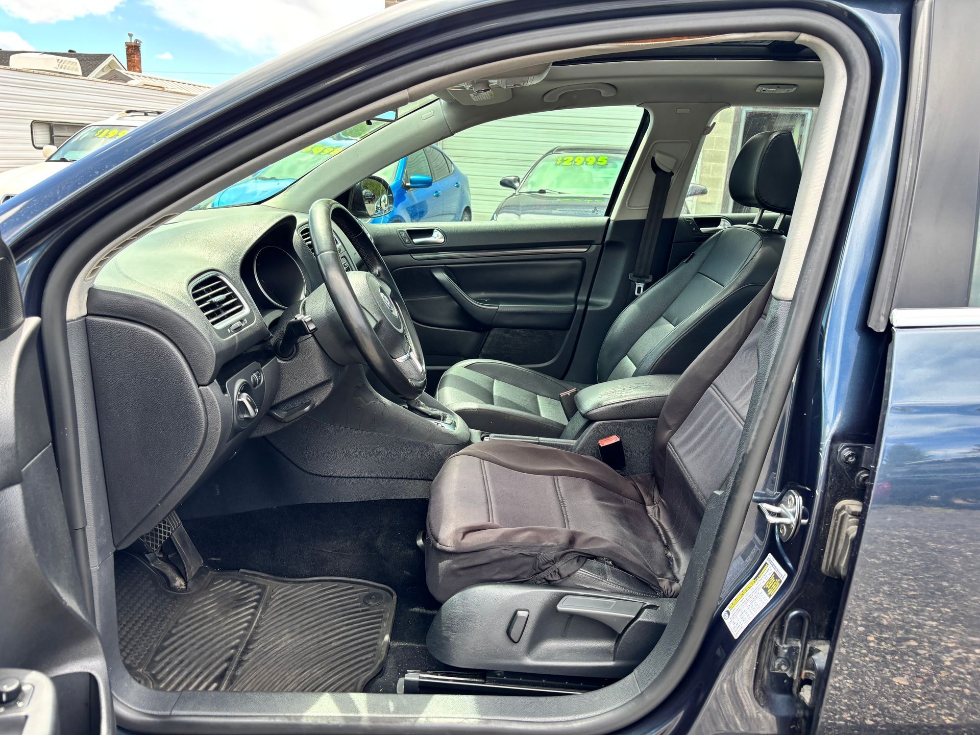 Open car door showing a black front seat with a worn cover and steering wheel inside a blue vehicle