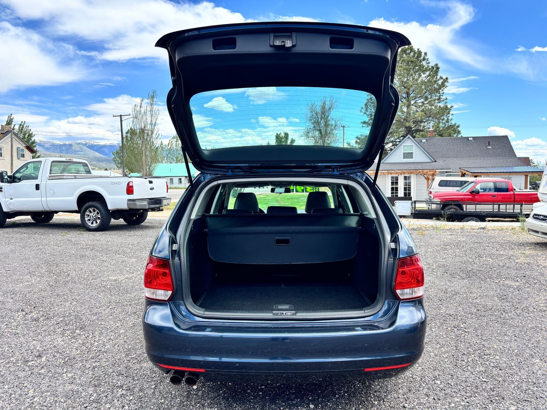 Open hatchback of a dark blue car in a gravel lot with trucks and houses in the background