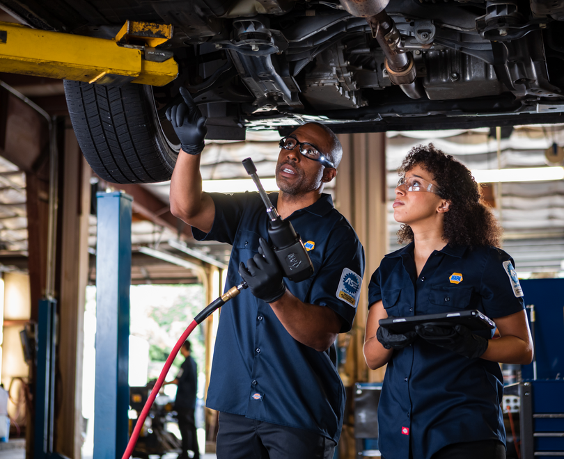 Two mechanics in blue uniforms inspect a vehicle underside in an auto repair shop.