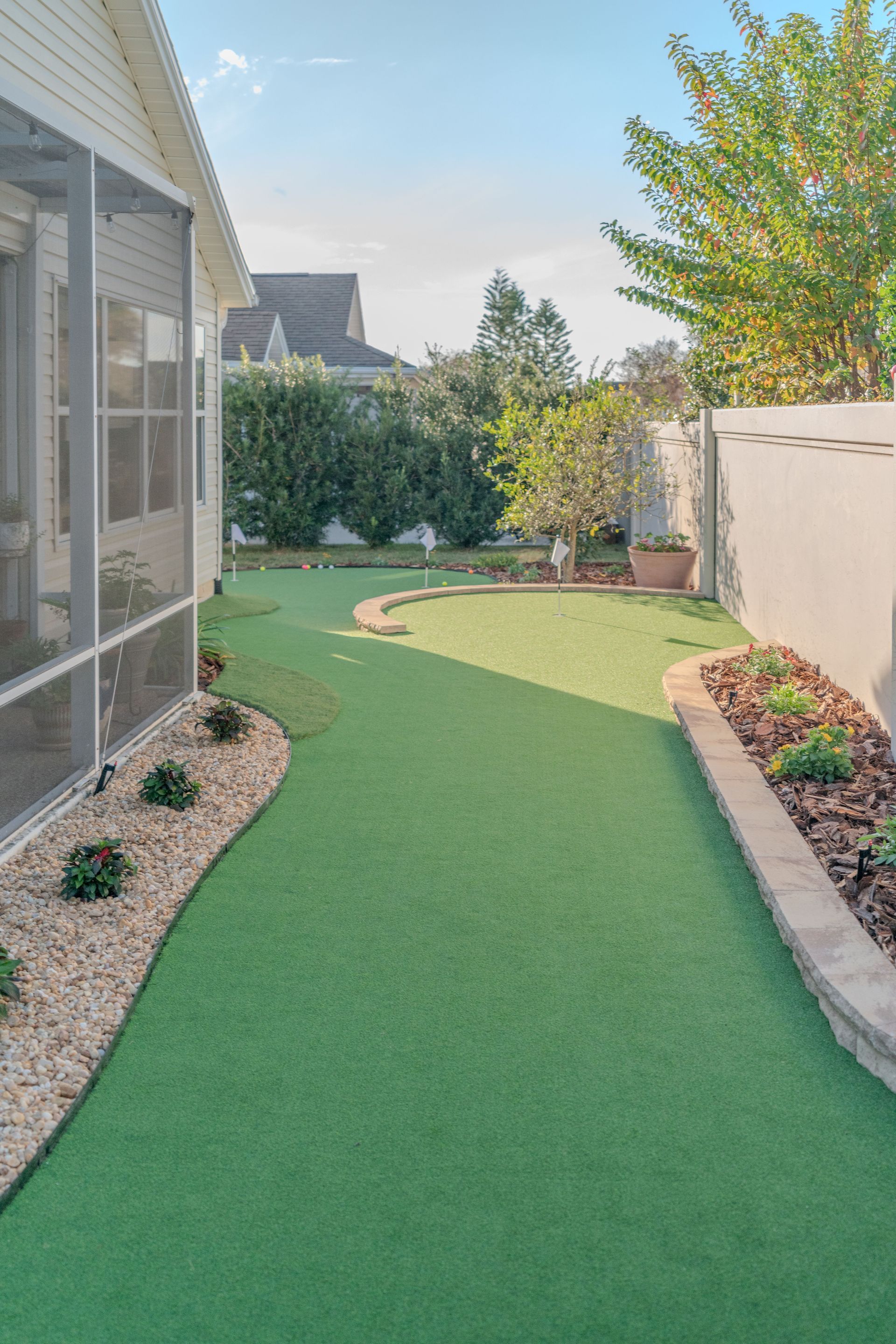 Narrow backyard with green artificial turf, stone border, shrubs, and a house wall along the left side.