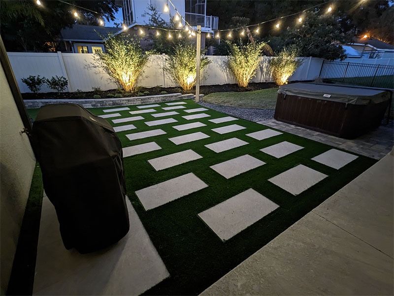 Backyard patio with string lights, illuminated trees, and white stepping stones on artificial grass around a hot tub