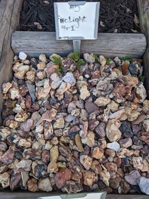 Small pile of multicolored gravel stones beneath a labeled sign in a garden bed