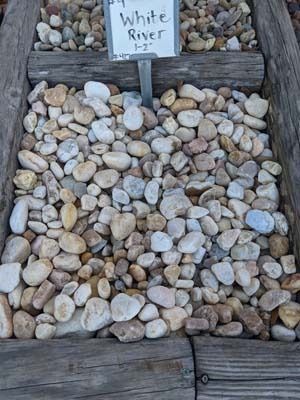 Box of smooth white and tan pebbles labeled “White River” on a wooden display shelf