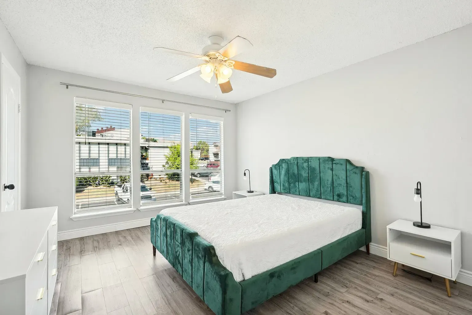 Bright bedroom with teal upholstered bed, white dressers, and large windows with blinds.