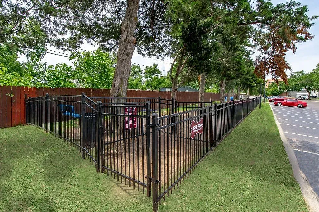Fenced dog park at an apartment community with trees and a bench.