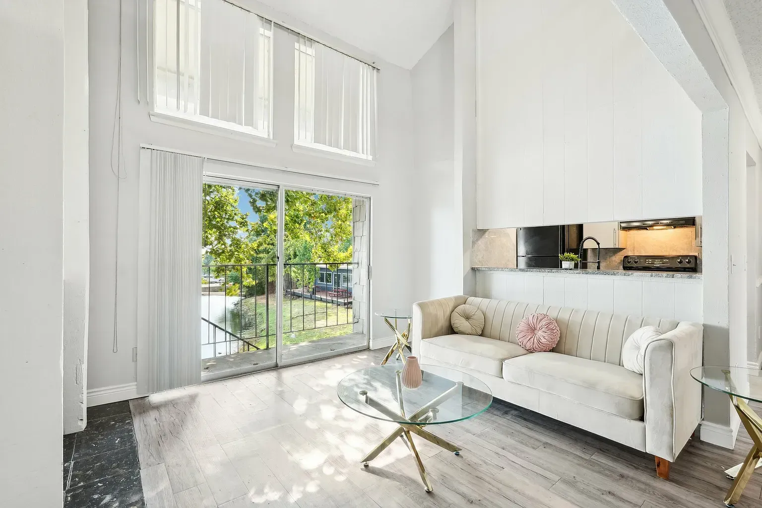 Bright living room with white walls, sofa, glass coffee table, and sliding glass door to a balcony.