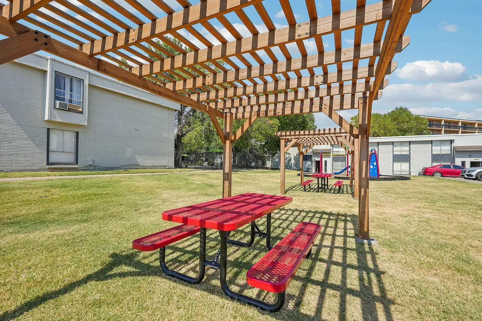 Wooden pergolas over red picnic tables on a grassy courtyard with a playground in the background.