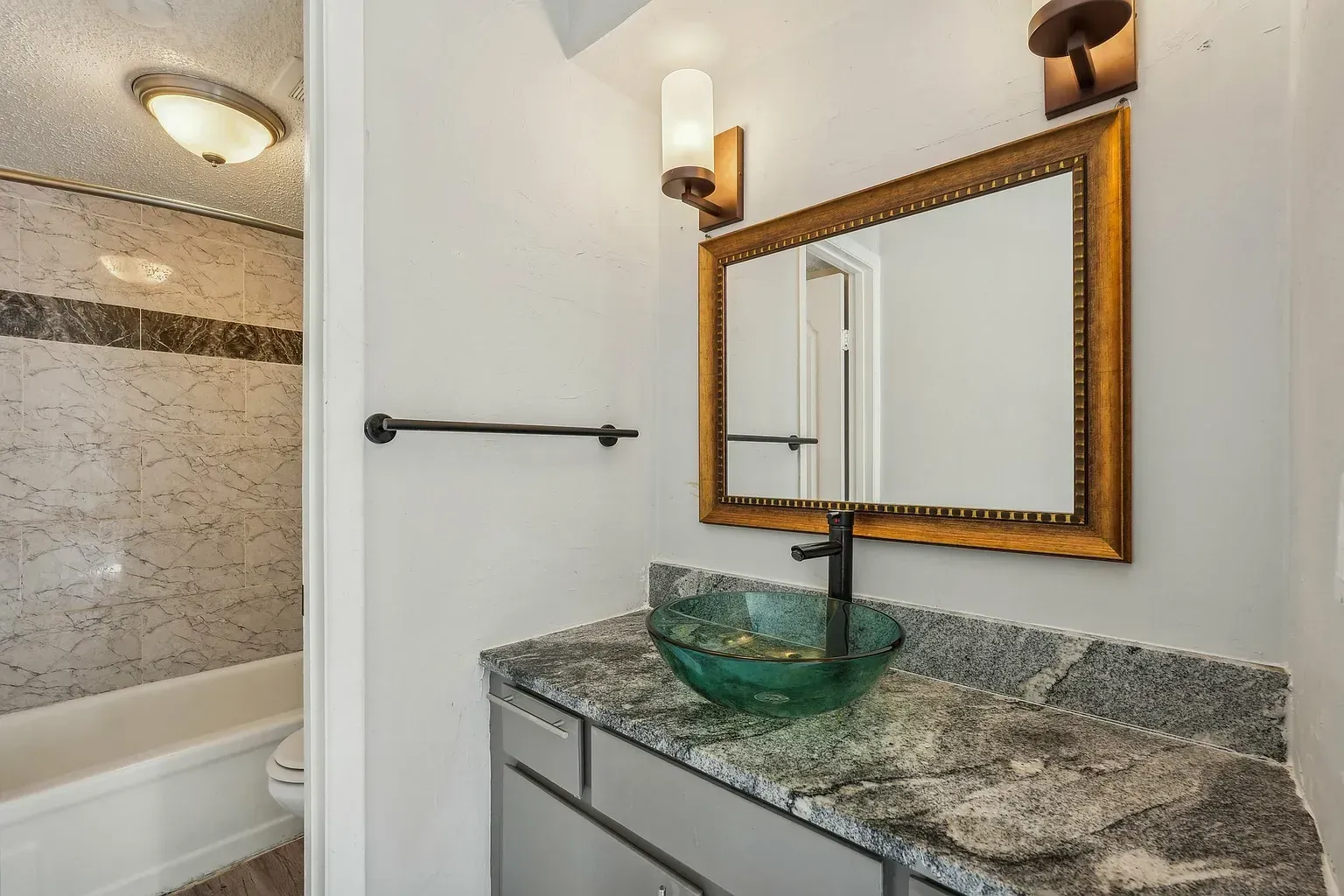 Bathroom vanity with a green glass vessel sink, granite countertop, and framed mirror; shower in the background.