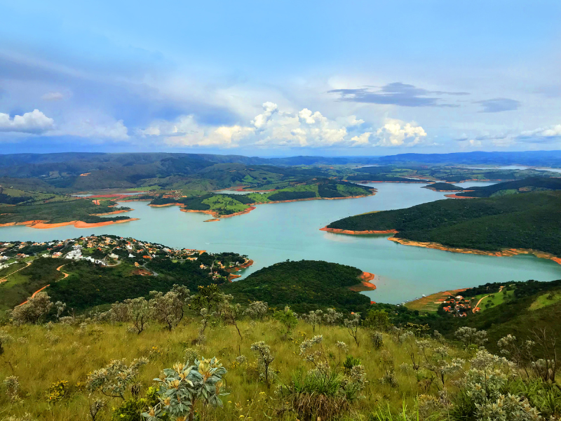 Vista panorâmica de um lago e colinas verdejantes sob um céu azul nublado, a partir de uma encosta gramada.