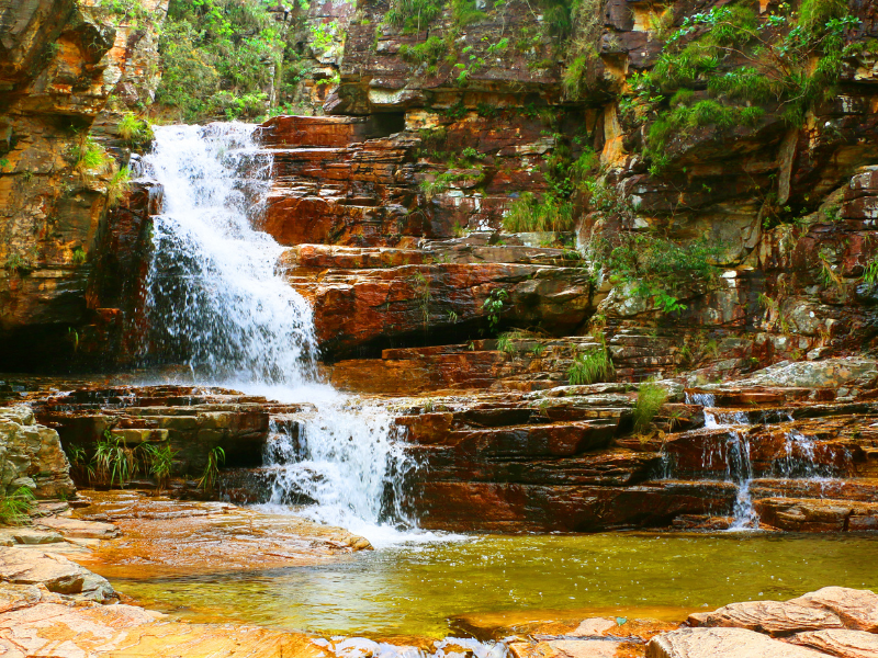 Cachoeira em degraus que deságua sobre rochas vermelhas em uma piscina verdejante na floresta.