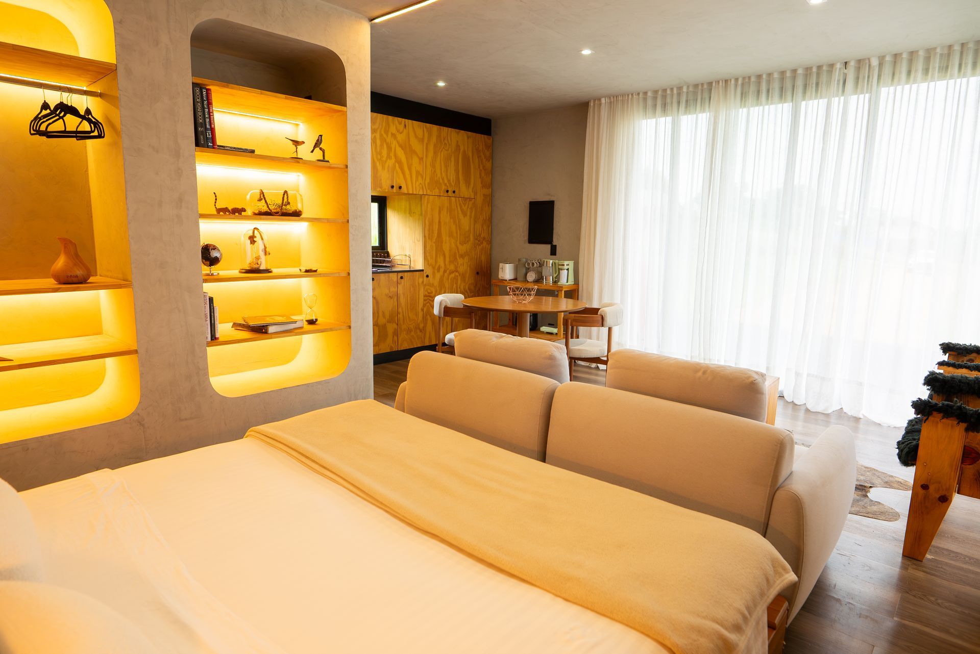 Quarto moderno e aconchegante com roupa de cama bege, prateleiras embutidas e cortinas translúcidas em janelas amplas.