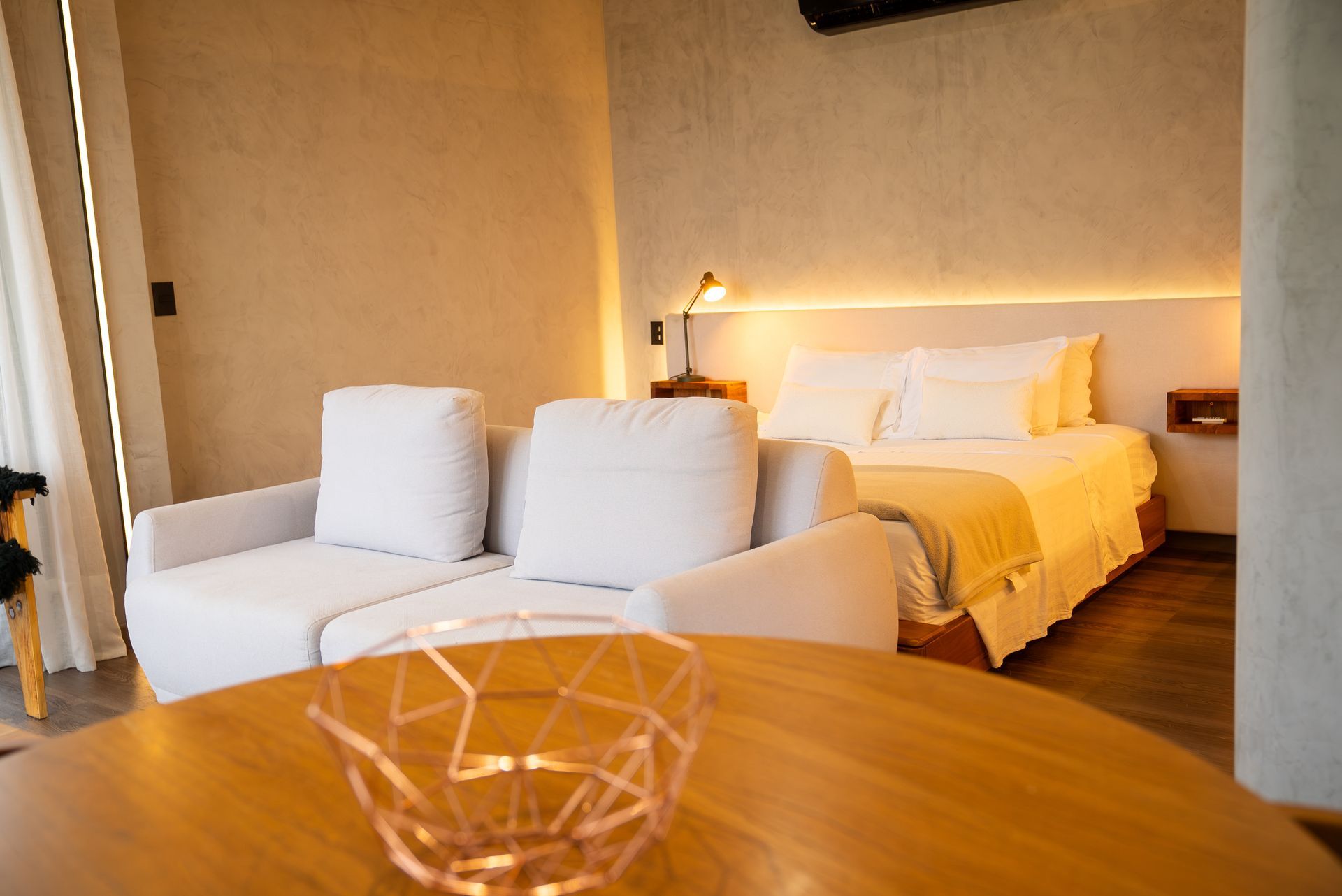 Quarto de hotel aconchegante com sofá branco, cama, iluminação suave de abajur e uma mesa de madeira em primeiro plano.