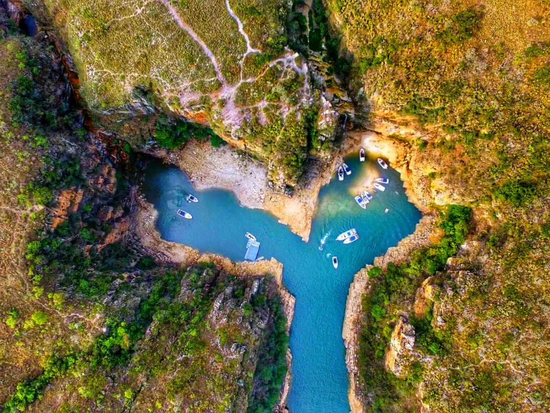 Vista aérea de uma piscina natural azul em um desfiladeiro rochoso em forma de Y, cercada por vegetação verde-acastanhada.