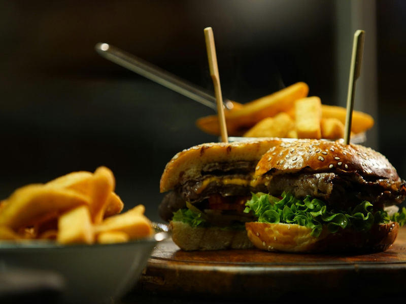 Hambúrguer gourmet com batatas fritas sobre uma mesa escura, com pauzinhos e uma tigela de batatas fritas ao fundo.