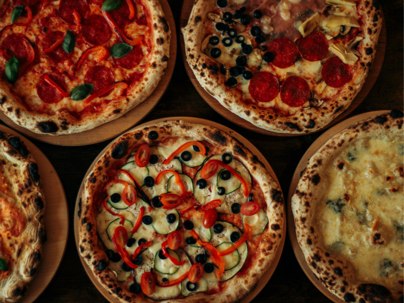 Pizzas variadas com diversos ingredientes, incluindo pepperoni, azeitonas e vegetais, dispostas sobre uma superfície escura.