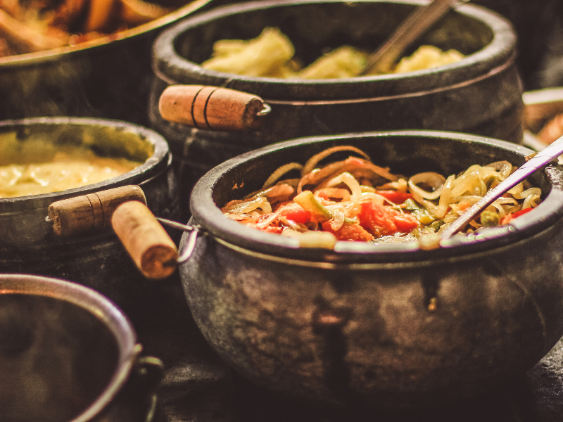 Pratos de macarrão cozidos em panelas rústicas pretas com cabos de madeira, servidos em estilo bufê.