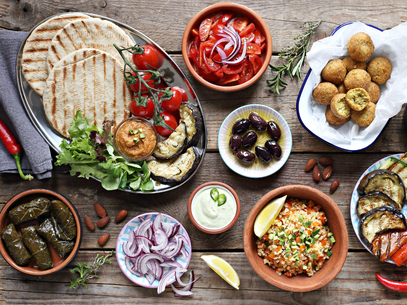 Seleção de aperitivos mediterrâneos com wraps, legumes grelhados, molhos, azeitonas e salada sobre uma mesa escura.